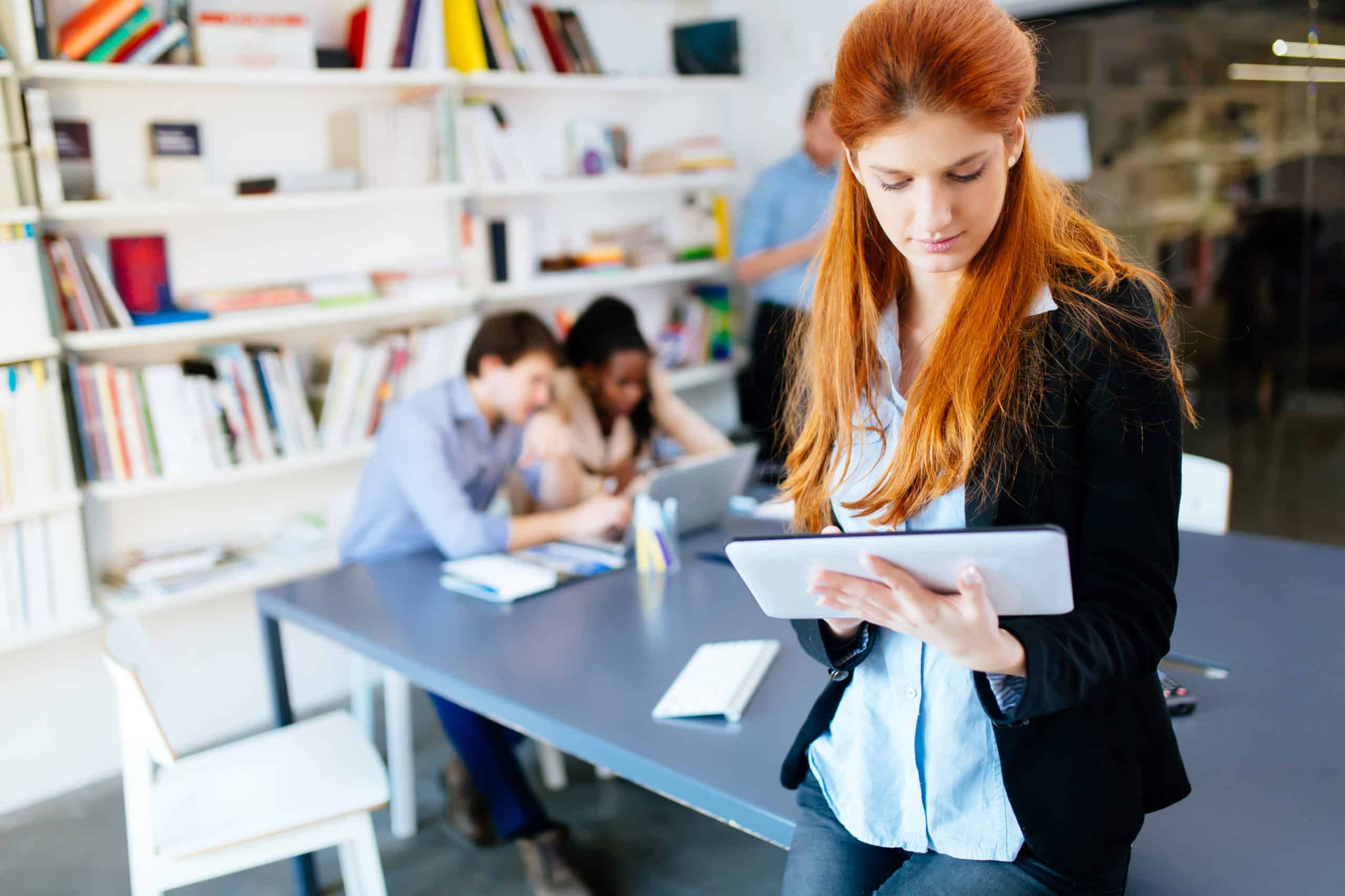 Redhead businesswoman holding a tablet in an office with coworkers communicating in the background