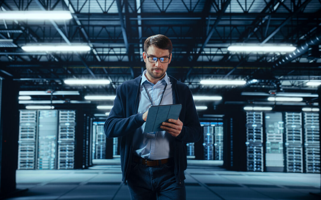 Male IT Specialist Walks Between Row of Operational Server Racks in Data Center. Engineer Uses Tablet Computer for Maintenance. Concept for Cloud Computing, Artificial Intelligence, Cybersecurity